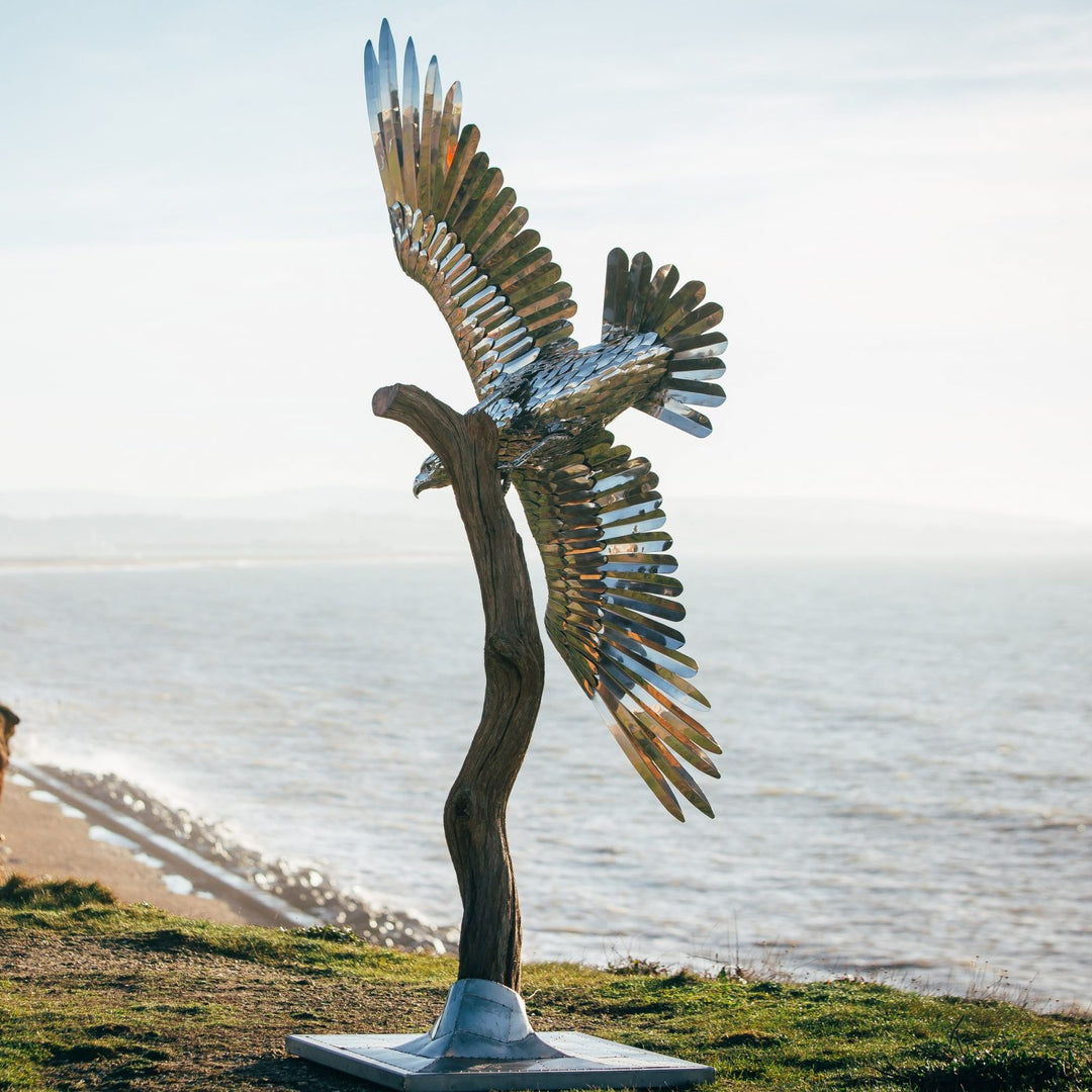 ‘Golden Eagle’ by Michael Turner, a unique, one-of-one stainless steel wildlife sculpture depicting a magnificent golden eagle soaring with its powerful wings fully extended. Handcrafted from polished stainless steel, the sculpture features individually shaped metallic feathers, intricately detailed plumage, a sharply curved beak and striking eyes. Finished in reflective silver with subtle gold tones from the surrounding light, the eagle is mounted on a tall, weathered tree trunk rising from a square metal 