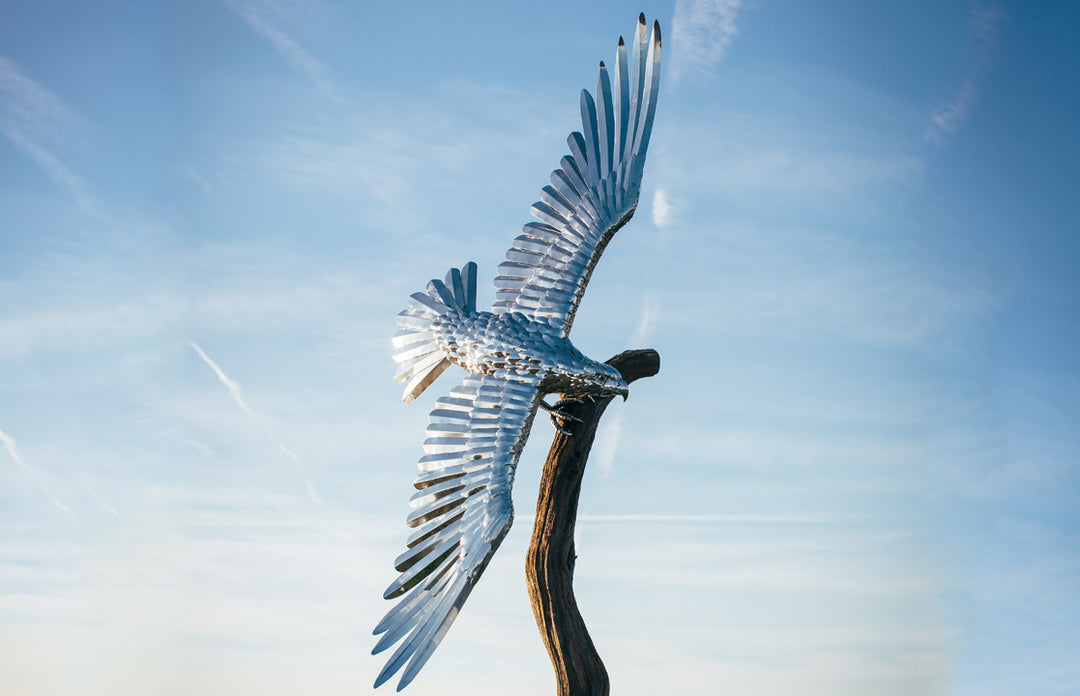 ‘Golden Eagle’ by Michael Turner, a unique, one-of-one stainless steel wildlife sculpture depicting a magnificent golden eagle soaring with its powerful wings fully extended. Handcrafted from polished stainless steel, the sculpture features individually shaped metallic feathers, intricately detailed plumage, a sharply curved beak and striking eyes. Finished in reflective silver with subtle gold tones from the surrounding light, the eagle is mounted on a tall, weathered tree trunk rising from a square metal 