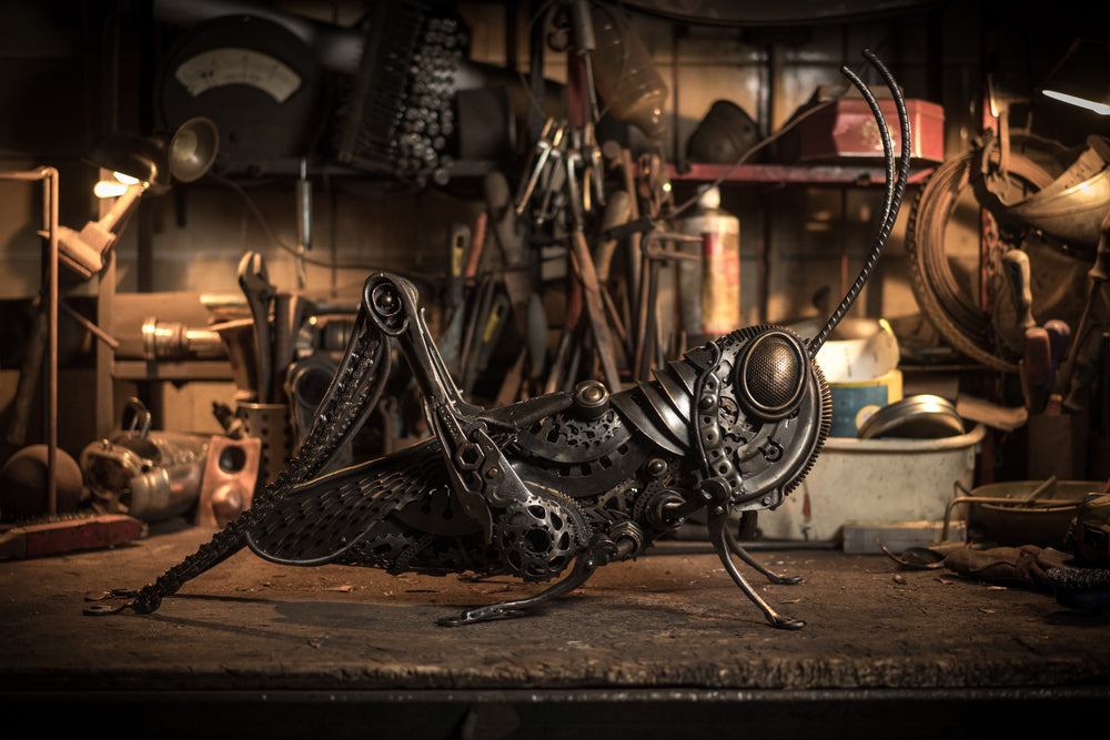 Mechanical grasshopper sculpture crafted from reclaimed metal and gears, displayed on a rustic workbench.
