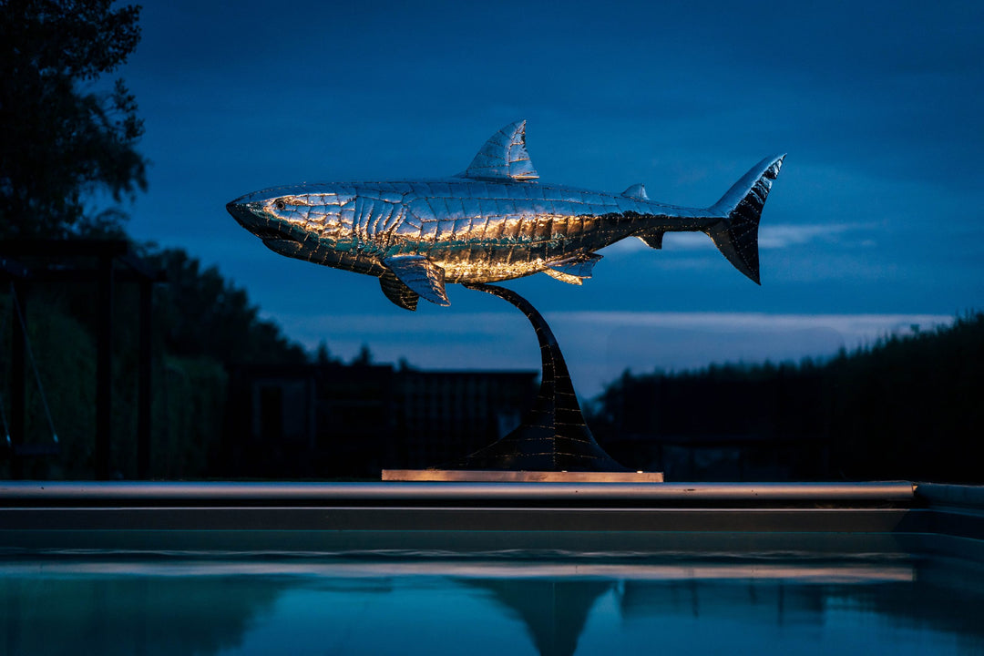 ‘Great White’ by Michael Turner, a unique, one-of-one stainless steel marine wildlife sculpture depicting a powerful great white shark swimming with its mouth partially open, exposing rows of sharp triangular teeth. Handcrafted from intricately shaped and welded stainless steel, the sculpture features a polished silver finish, detailed metallic skin, distinctive dorsal and pectoral fins and a sweeping tail. Mounted on a curved black metal support with a rectangular base, this large-scale contemporary sculpt