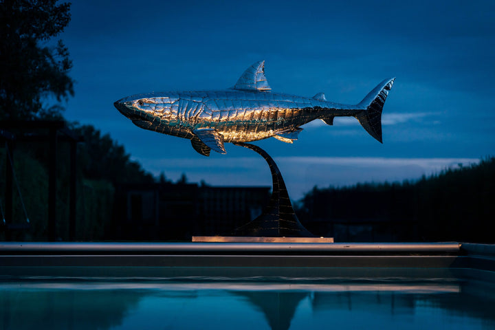 ‘Great White’ by Michael Turner, a unique, one-of-one stainless steel marine wildlife sculpture depicting a powerful great white shark swimming with its mouth partially open, exposing rows of sharp triangular teeth. Handcrafted from intricately shaped and welded stainless steel, the sculpture features a polished silver finish, detailed metallic skin, distinctive dorsal and pectoral fins and a sweeping tail. Mounted on a curved black metal support with a rectangular base, this large-scale contemporary sculpt