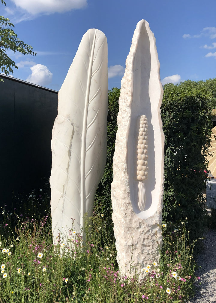 ‘Guardians’ by Nicolas Moreton MRSS, a pair of unique contemporary botanical stone sculptures carved from pale white Portuguese Marble. The first sculpture features a tall, upright leaf with a smooth surface and finely carved central vein and branching leaf veins. The second features an elongated, pointed form with rough natural stone edges, a deeply recessed smooth centre and a raised, textured seed-like structure. Both sculptures stand 260cm tall and are photographed together in a landscaped garden surrou