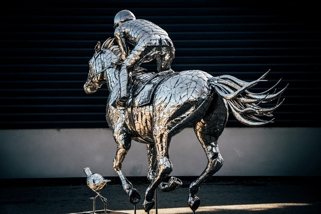 ‘Racehorse & Jockey - A Dynamic Force’ by Michael Turner, a unique, one-of-one stainless steel equestrian sculpture depicting a powerful thoroughbred racehorse galloping at full speed with its jockey leaning forward in a racing position. Handcrafted from hundreds of individually shaped and welded marine-grade stainless steel pieces, the sculpture features a polished silver finish, detailed muscular anatomy, flowing mane and tail, and a helmeted jockey holding the reins. Accompanied by a small running bird, 