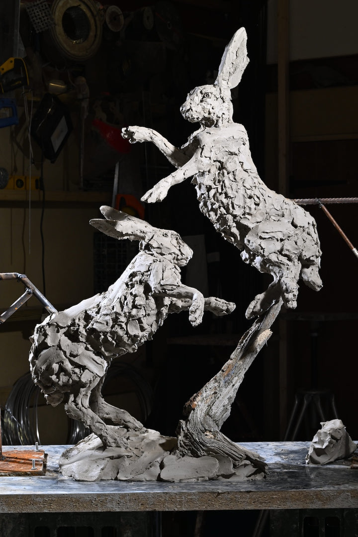 ‘Boxing Hares’ by Hamish Mackie, a limited-edition bronze wildlife sculpture depicting two hares leaping and boxing in mid-air during their characteristic springtime courtship behaviour. The dynamic composition captures their extended limbs, twisting bodies and expressive movement, with richly textured surfaces that convey the energy of the original hand-modelled clay. Photograph shows the original clay sculpture prior to bronze casting.