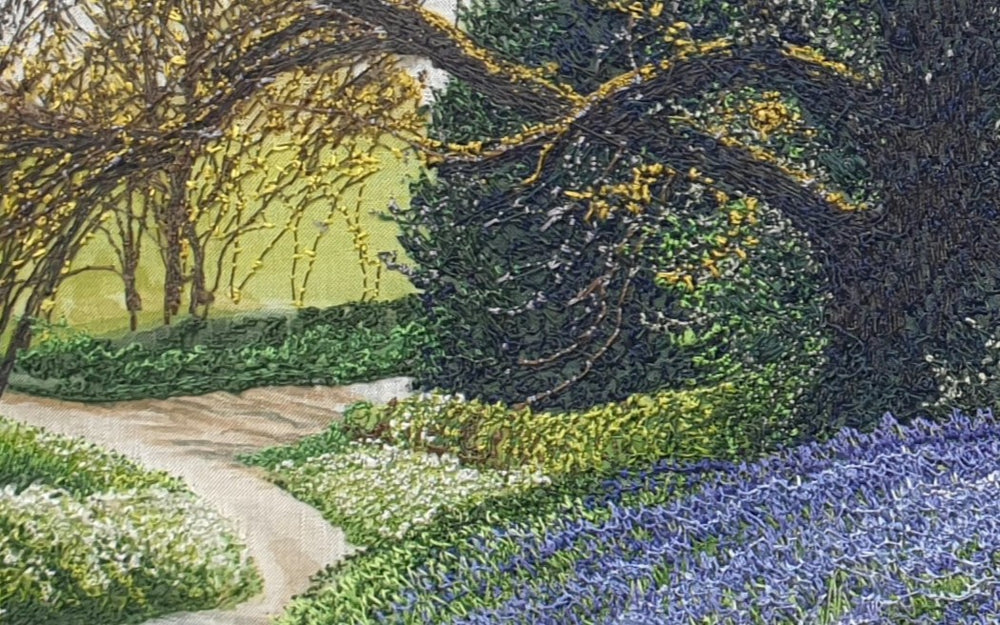 Textile artwork depicting a bluebell-covered landscape with a large oak tree and winding path, created with freehand machine embroidery on painted silk.