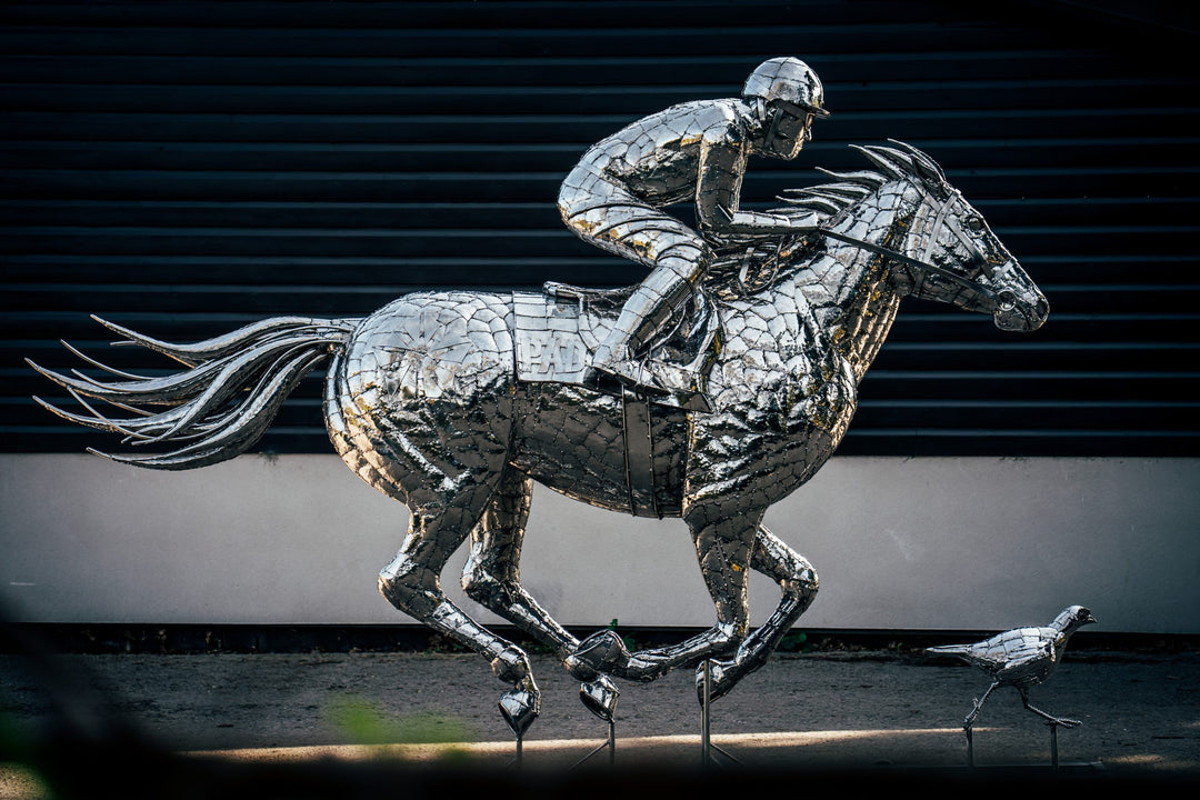 ‘Racehorse & Jockey - A Dynamic Force’ by Michael Turner, a unique, one-of-one stainless steel equestrian sculpture depicting a powerful thoroughbred racehorse galloping at full speed with its jockey leaning forward in a racing position. Handcrafted from hundreds of individually shaped and welded marine-grade stainless steel pieces, the sculpture features a polished silver finish, detailed muscular anatomy, flowing mane and tail, and a helmeted jockey holding the reins. Accompanied by a small running bird, 