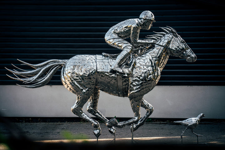 ‘Racehorse & Jockey - A Dynamic Force’ by Michael Turner, a unique, one-of-one stainless steel equestrian sculpture depicting a powerful thoroughbred racehorse galloping at full speed with its jockey leaning forward in a racing position. Handcrafted from hundreds of individually shaped and welded marine-grade stainless steel pieces, the sculpture features a polished silver finish, detailed muscular anatomy, flowing mane and tail, and a helmeted jockey holding the reins. Accompanied by a small running bird, 
