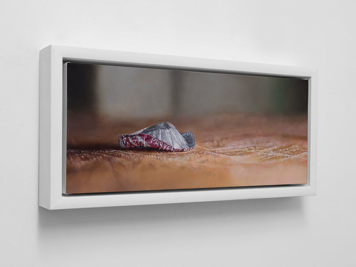Simon Taylor, How The End Always Is, oil on canvas, 17 x 41cm – photorealist painting of a crumpled red and silver bottle cap resting on a warm brown wooden surface with shallow depth of field and soft blurred background.