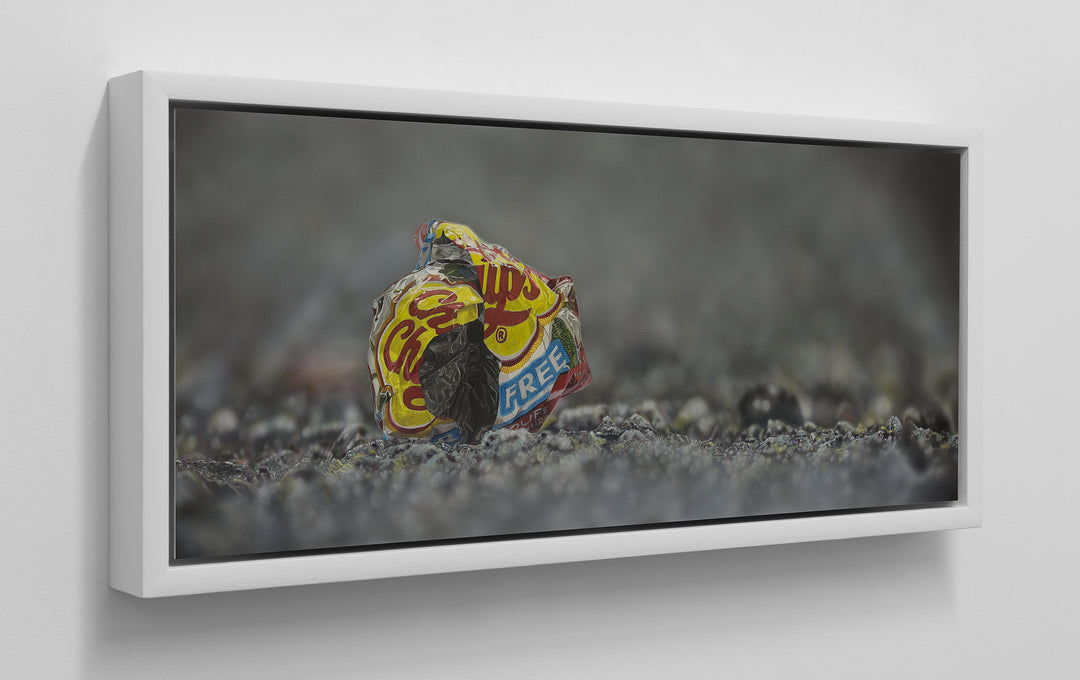 Simon Taylor, I Gave You My Heart (Original), oil and acrylic on aluminium, 25 x 60cm – photorealist painting of a crumpled red and yellow crisp wrapper resting on dark gravel against a soft grey background.