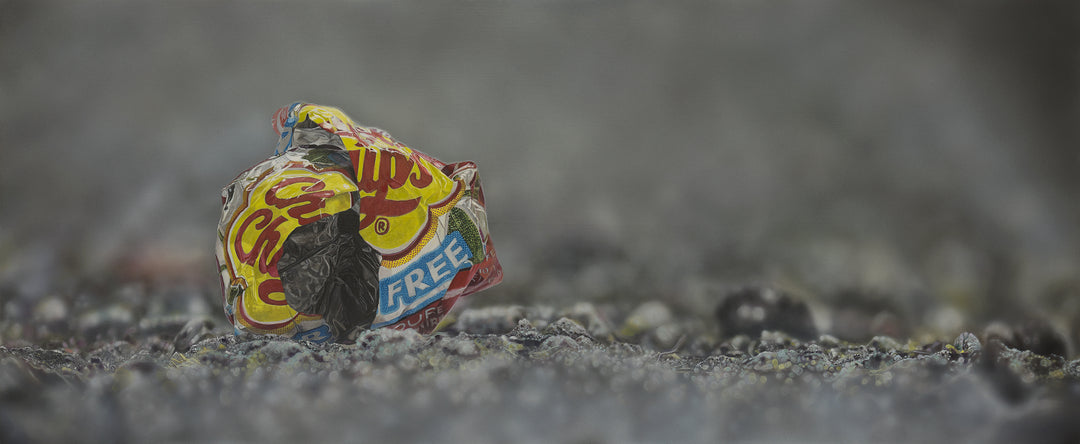Simon Taylor, I Gave You My Heart (Original), oil and acrylic on aluminium, 25 x 60cm – photorealist painting of a crumpled red and yellow crisp wrapper resting on dark gravel against a soft grey background.
