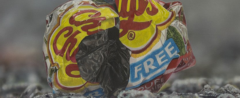 Simon Taylor, I Gave You My Heart (Original), oil and acrylic on aluminium, 25 x 60cm – photorealist painting of a crumpled red and yellow crisp wrapper resting on dark gravel against a soft grey background.