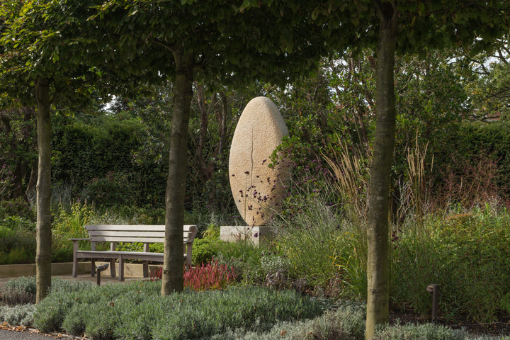 ‘New Beginnings’ by Nicolas Moreton MRSS, a unique commissioned contemporary abstract stone sculpture carved from Ancaster Weatherbed Limestone, featuring a tall, upright oval form with a textured surface and a narrow, irregular vertical opening illuminated by warm white LED light. The sculpture stands on a Portland limestone base in a landscaped garden at dusk, with its illuminated centre contrasting against the dark surrounding trees, purple garden lighting and evening sky.