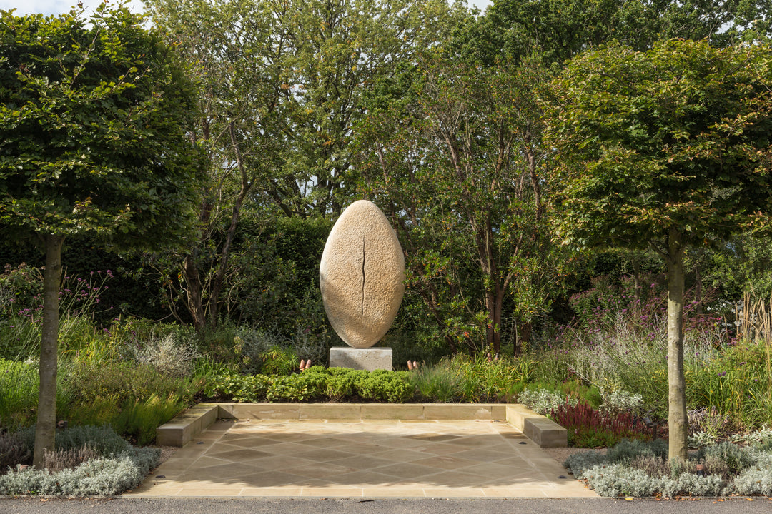 ‘New Beginnings’ by Nicolas Moreton MRSS, a unique commissioned contemporary abstract stone sculpture carved from Ancaster Weatherbed Limestone, featuring a tall, upright oval form with a textured surface and a narrow, irregular vertical opening illuminated by warm white LED light. The sculpture stands on a Portland limestone base in a landscaped garden at dusk, with its illuminated centre contrasting against the dark surrounding trees, purple garden lighting and evening sky.