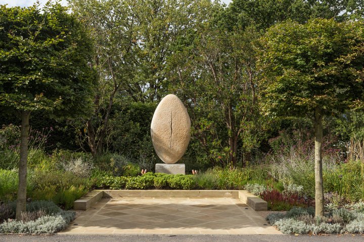 ‘New Beginnings’ by Nicolas Moreton MRSS, a unique commissioned contemporary abstract stone sculpture carved from Ancaster Weatherbed Limestone, featuring a tall, upright oval form with a textured surface and a narrow, irregular vertical opening illuminated by warm white LED light. The sculpture stands on a Portland limestone base in a landscaped garden at dusk, with its illuminated centre contrasting against the dark surrounding trees, purple garden lighting and evening sky.