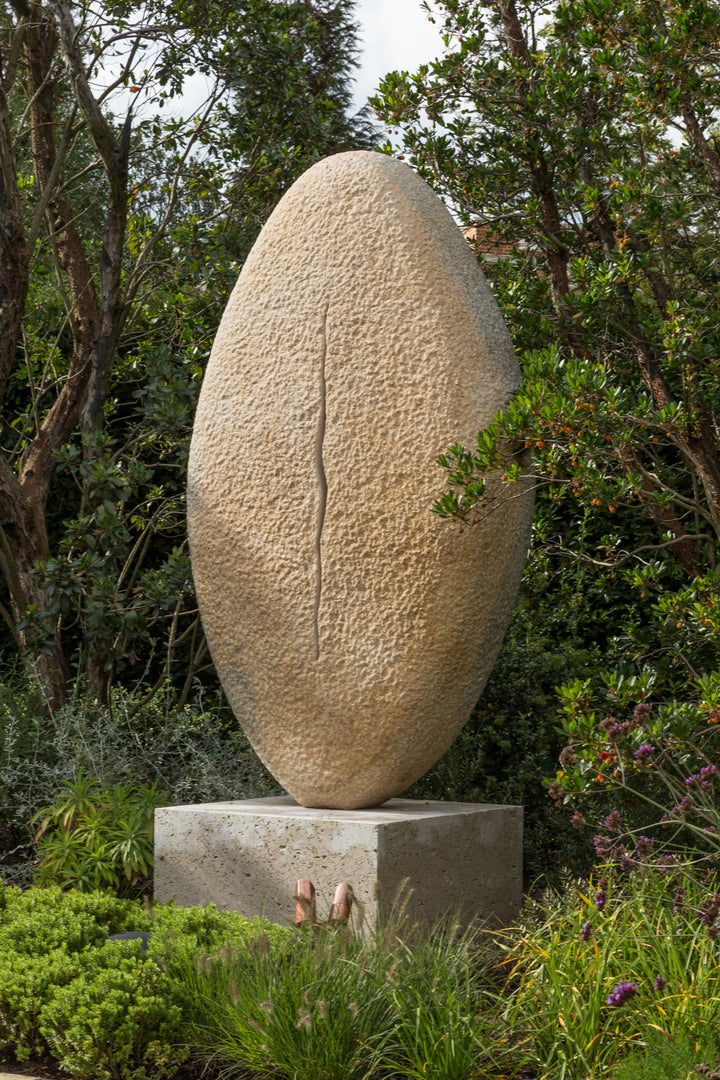 ‘New Beginnings’ by Nicolas Moreton MRSS, a unique commissioned contemporary abstract stone sculpture carved from Ancaster Weatherbed Limestone, featuring a tall, upright oval form with a textured surface and a narrow, irregular vertical opening illuminated by warm white LED light. The sculpture stands on a Portland limestone base in a landscaped garden at dusk, with its illuminated centre contrasting against the dark surrounding trees, purple garden lighting and evening sky.