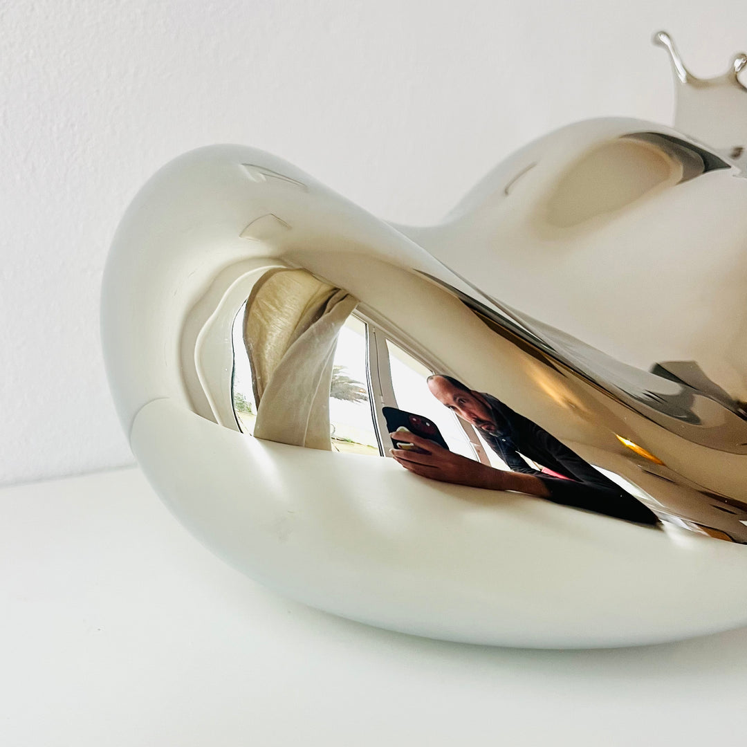 Polished stainless steel sculpture by Yoni Alter capturing the elegant moment of a liquid droplet mid-bounce. The reflective form twists fluidly to the left, with raised crown-like peaks suggesting impact and motion. A sculptural meditation on impermanence and form, grounded by a hidden counterweight for stability.