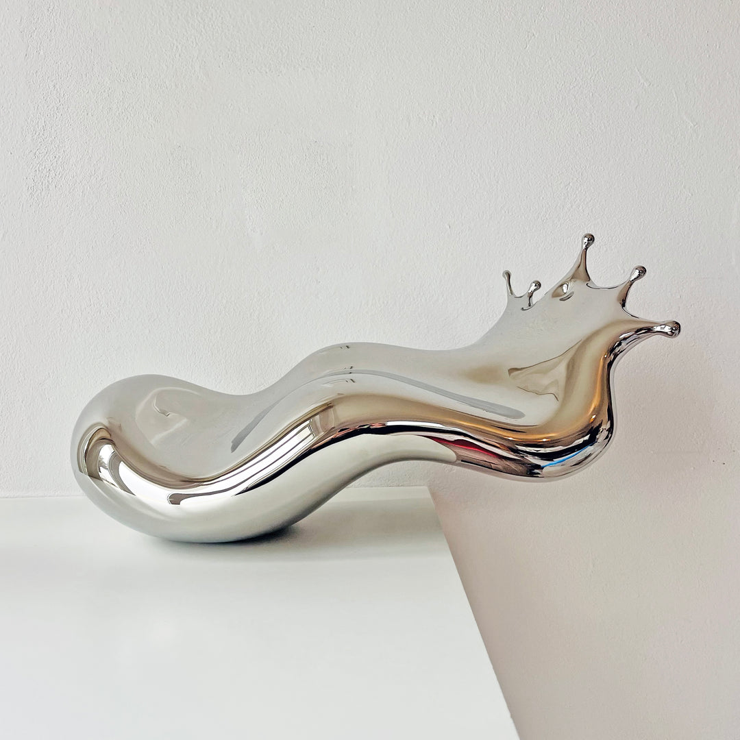 Polished stainless steel sculpture by Yoni Alter capturing the elegant moment of a liquid droplet mid-bounce. The reflective form twists fluidly to the left, with raised crown-like peaks suggesting impact and motion. A sculptural meditation on impermanence and form, grounded by a hidden counterweight for stability.