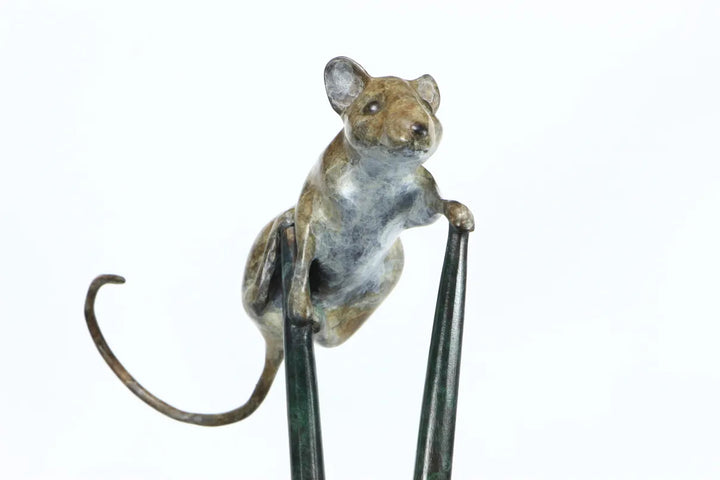 Bronze sculpture of a curious mouse perched high atop two crossed stilts, tail arched playfully behind, poised in a moment of bold exploration and balance.