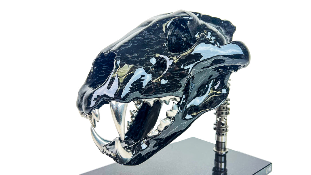 A striking, polished carbon fibre lion skull sculpture by Alastair Gibson, crafted with Mokume-style carbon layup for a swirling, chaotic texture. The jaw is fitted with gleaming bismuth alloy teeth. Mounted on a genuine F1 gearbox selector barrel atop a granite base, the work merges natural form with elite motorsport engineering.