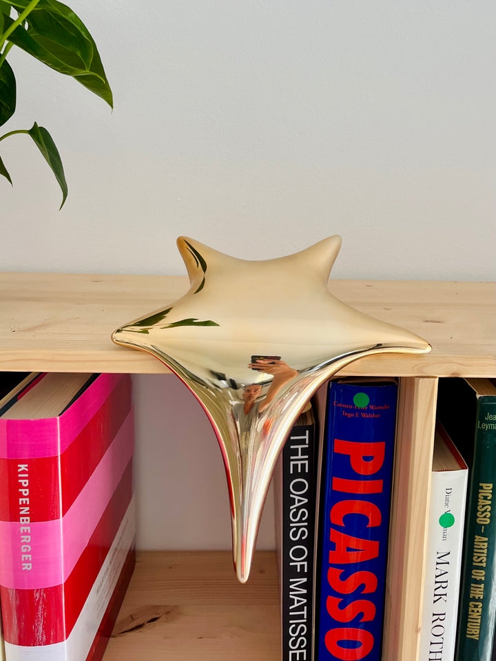 Polished gold-toned stainless steel sculpture in the form of a stylised shooting star, with a fluid, elongated spike melting over the edge of a white plinth or shelf.