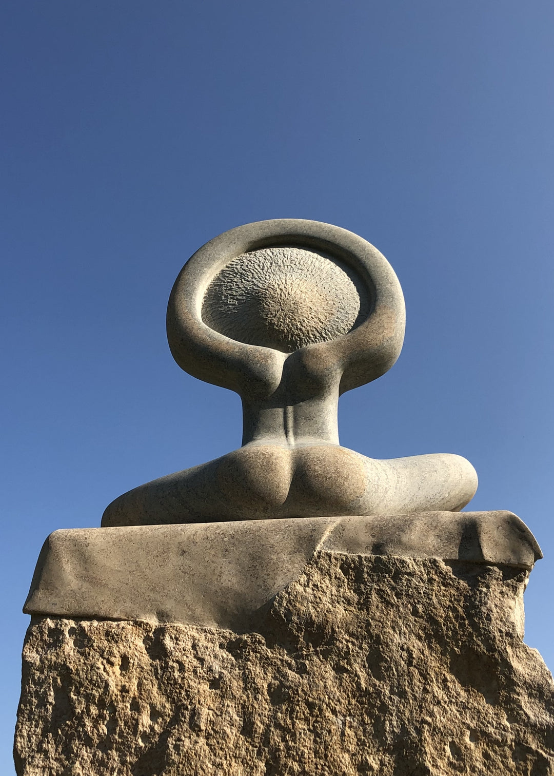 ‘Sunrise’ by Nicolas Moreton MRSS, a unique contemporary figurative stone sculpture carved from Ancaster Weatherbed Limestone, depicting a stylised seated female figure with crossed legs and raised arms forming a circular halo-like shape above her head. The figure features a delicately carved face, textured hair and smooth, rounded body, emerging from a large rectangular limestone block with a rough natural surface and flowing carved folds. Finished in warm cream, pale beige and natural limestone tones, the