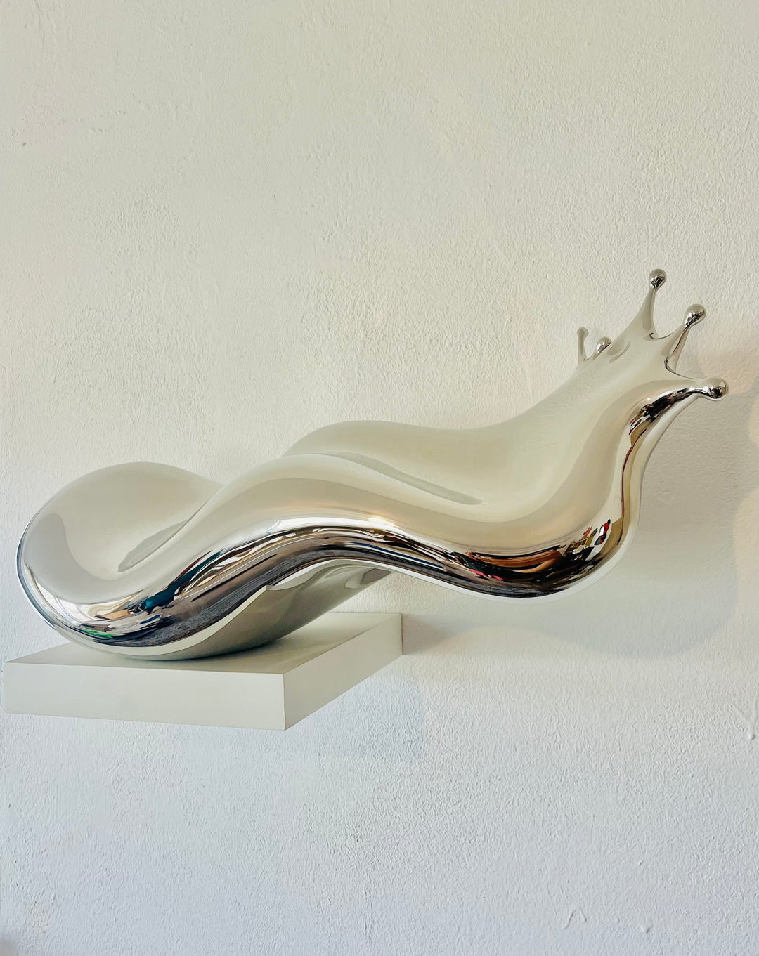 Polished stainless steel sculpture by Yoni Alter capturing the elegant moment of a liquid droplet mid-bounce. The reflective form twists fluidly to the left, with raised crown-like peaks suggesting impact and motion. A sculptural meditation on impermanence and form, grounded by a hidden counterweight for stability.