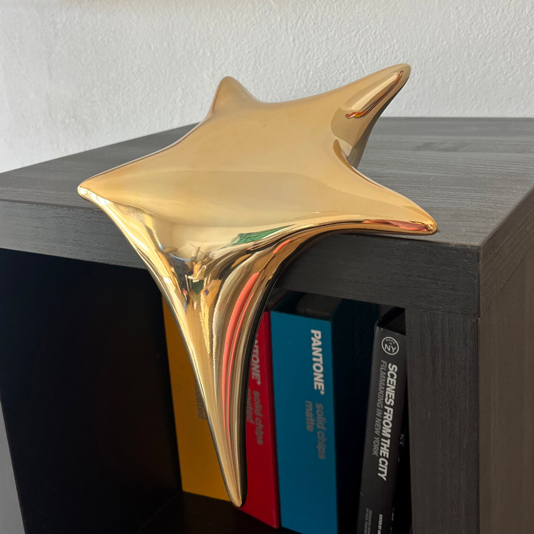 Polished gold-toned stainless steel sculpture in the form of a stylised shooting star, with a fluid, elongated spike melting over the edge of a white plinth or shelf.