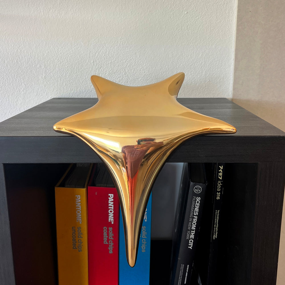 Polished gold-toned stainless steel sculpture in the form of a stylised shooting star, with a fluid, elongated spike melting over the edge of a white plinth or shelf.