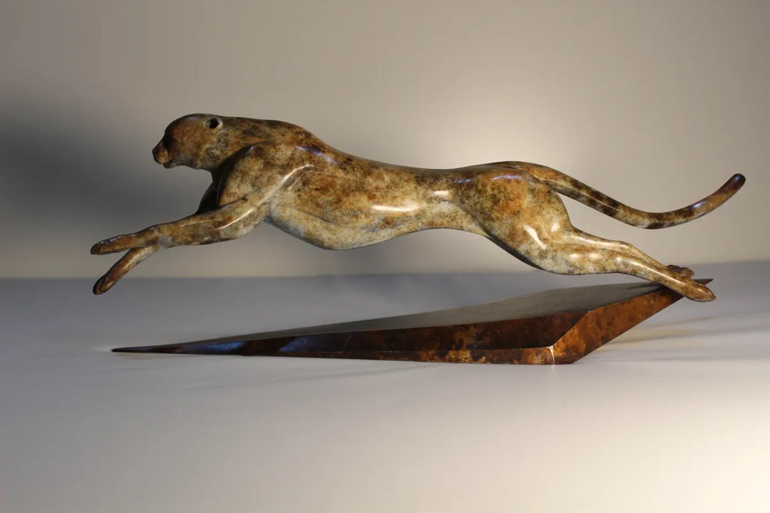 Sleek bronze sculpture of a leaping cheetah with a rich mottled patina, balanced dynamically on a tapered triangular bronze base. The form is elegant and streamlined, capturing a sense of speed and fluid motion.