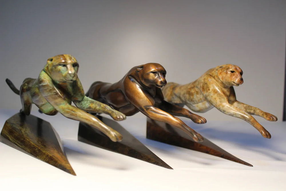 Sleek bronze sculpture of a leaping cheetah with a rich mottled patina, balanced dynamically on a tapered triangular bronze base. The form is elegant and streamlined, capturing a sense of speed and fluid motion.