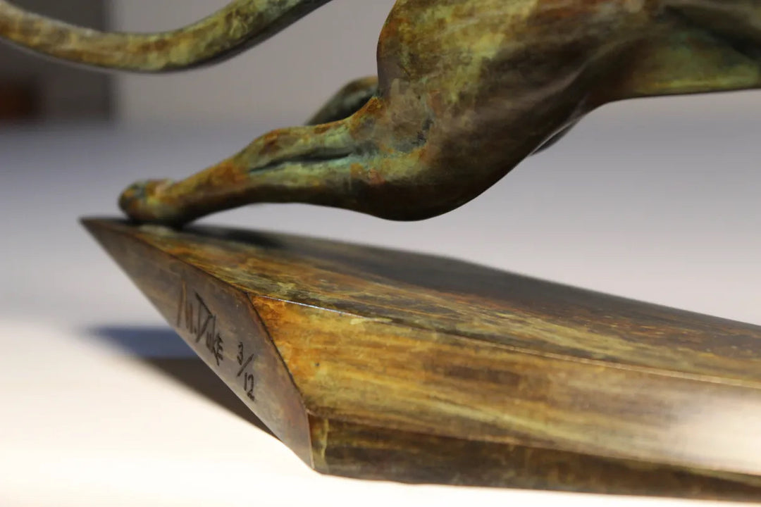 Sleek bronze sculpture of a leaping cheetah with a rich mottled patina, balanced dynamically on a tapered triangular bronze base. The form is elegant and streamlined, capturing a sense of speed and fluid motion.