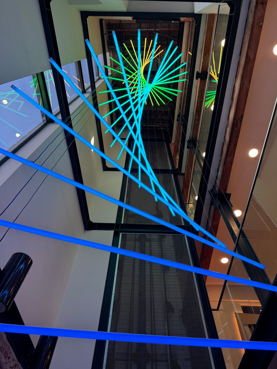 Suspended spiral light installation composed of intersecting illuminated linear elements arranged around a vertical stairwell void, with coloured light bars radiating and twisting through space to echo the geometry and movement of the surrounding architecture, creating layered reflections and depth within the entrance atrium.