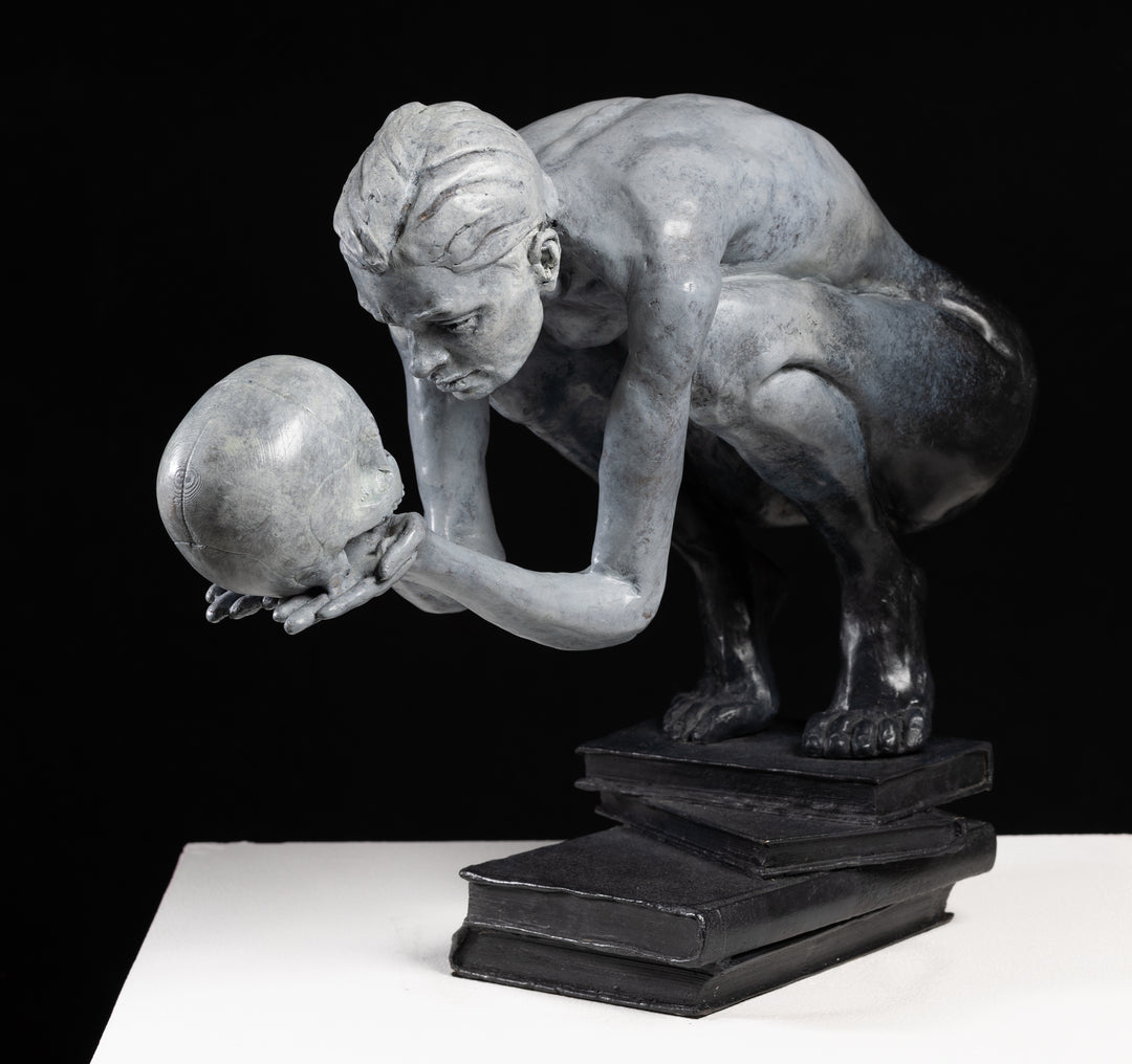 Teresa Wells’ The Prophecy (2024), limited edition bronze sculpture depicting a crouched, nude figure balanced atop stacked books, holding a human skull in both hands and gazing intently into its hollow eyes. Finished in a pale grey patina with darker shading to emphasise musculature and contours, the piece evokes themes of mortality, knowledge, and forewarning. At 30cm × 25cm × 49cm and 23.4kg, it combines fine anatomical detail, symbolic elements, and a contemporary reinterpretation of classical imagery.