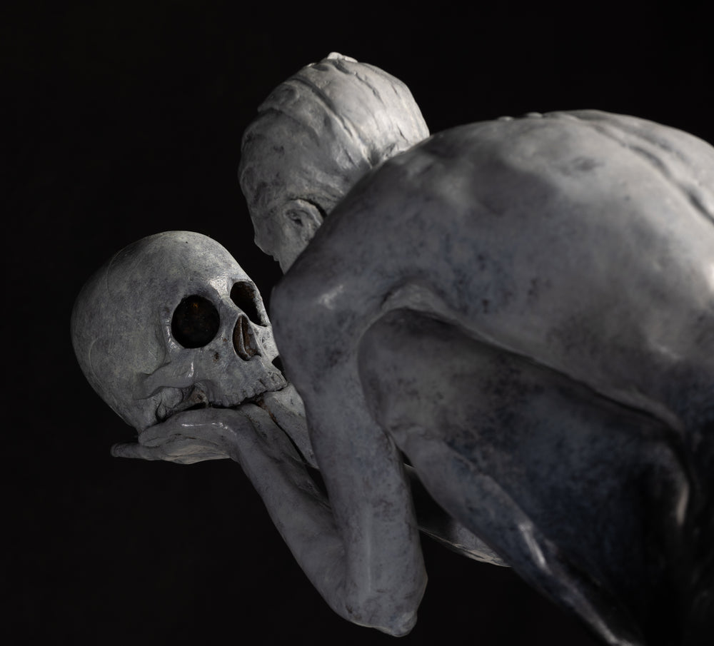 Teresa Wells’ The Prophecy (2024), limited edition bronze sculpture depicting a crouched, nude figure balanced atop stacked books, holding a human skull in both hands and gazing intently into its hollow eyes. Finished in a pale grey patina with darker shading to emphasise musculature and contours, the piece evokes themes of mortality, knowledge, and forewarning. At 30cm × 25cm × 49cm and 23.4kg, it combines fine anatomical detail, symbolic elements, and a contemporary reinterpretation of classical imagery.