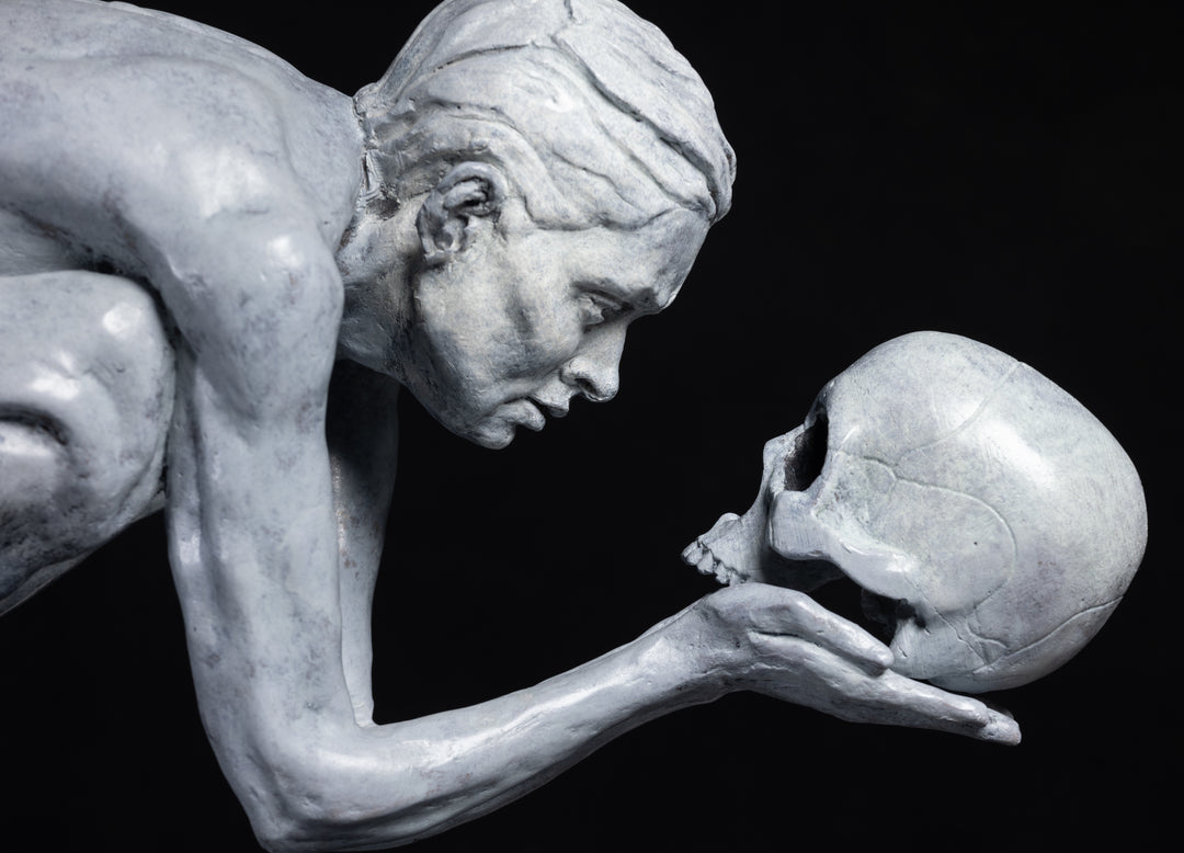 Teresa Wells’ The Prophecy (2024), limited edition bronze sculpture depicting a crouched, nude figure balanced atop stacked books, holding a human skull in both hands and gazing intently into its hollow eyes. Finished in a pale grey patina with darker shading to emphasise musculature and contours, the piece evokes themes of mortality, knowledge, and forewarning. At 30cm × 25cm × 49cm and 23.4kg, it combines fine anatomical detail, symbolic elements, and a contemporary reinterpretation of classical imagery.