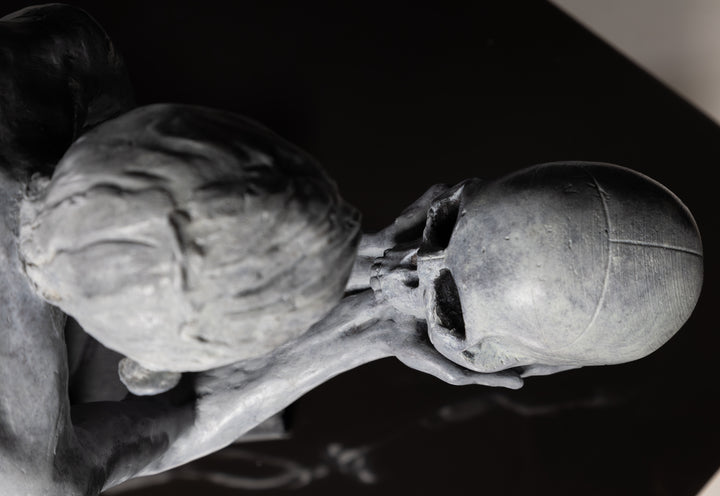 Teresa Wells’ The Prophecy (2024), limited edition bronze sculpture depicting a crouched, nude figure balanced atop stacked books, holding a human skull in both hands and gazing intently into its hollow eyes. Finished in a pale grey patina with darker shading to emphasise musculature and contours, the piece evokes themes of mortality, knowledge, and forewarning. At 30cm × 25cm × 49cm and 23.4kg, it combines fine anatomical detail, symbolic elements, and a contemporary reinterpretation of classical imagery.