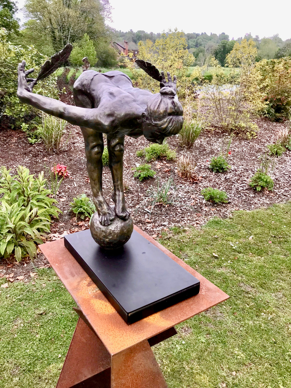 Ingenuity: Often thwarted by the shortsighted by Teresa Wells – a limited-edition bronze and steel sculpture depicting a poised, athletic female figure balanced on tiptoe atop a textured bronze sphere. The figure leans dramatically forward from the hips, arms extended behind and slightly upward, each hand delicately holding a detailed bronze feather. Muscular contours and tendons are rendered with lifelike precision, from the arched spine to the curled toes gripping the sphere. 