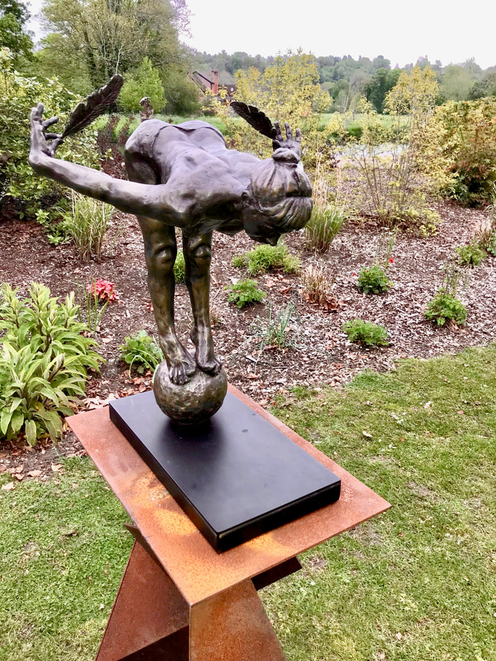 Ingenuity: Often thwarted by the shortsighted by Teresa Wells – a limited-edition bronze and steel sculpture depicting a poised, athletic female figure balanced on tiptoe atop a textured bronze sphere. The figure leans dramatically forward from the hips, arms extended behind and slightly upward, each hand delicately holding a detailed bronze feather. Muscular contours and tendons are rendered with lifelike precision, from the arched spine to the curled toes gripping the sphere. 