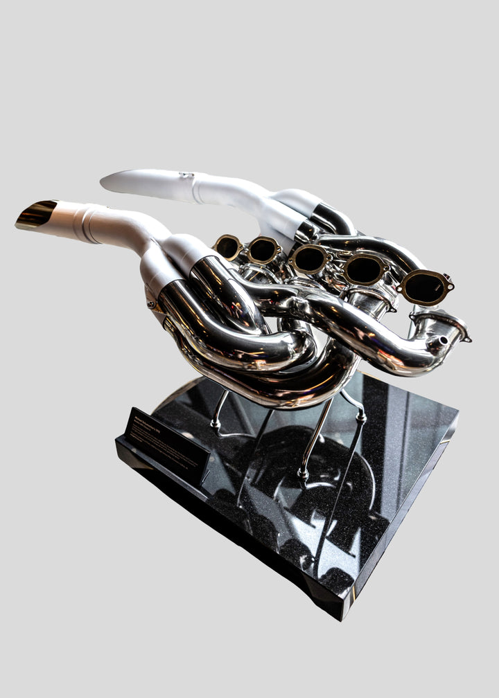 Highly polished sculptural artwork by Mike O’Connor crafted from the authentic race-used exhaust system of Michael Schumacher’s 1995 Benetton B195 Formula One car. The piece features flowing Inconel and stainless-steel pipes with mirror-finished chrome surfaces and 24-karat gold interior detailing, mounted on a sleek stand. A striking one-of-one collector’s piece 