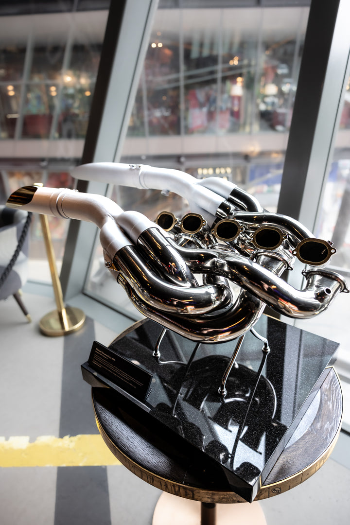 Highly polished sculptural artwork by Mike O’Connor crafted from the authentic race-used exhaust system of Michael Schumacher’s 1995 Benetton B195 Formula One car. The piece features flowing Inconel and stainless-steel pipes with mirror-finished chrome surfaces and 24-karat gold interior detailing, mounted on a sleek stand. A striking one-of-one collector’s piece 