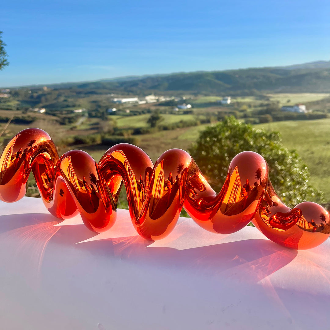 Freestanding sculptural artwork spelling ‘LOVE’ in a flowing, cursive form, finished in high-gloss orange-tinted stainless steel.