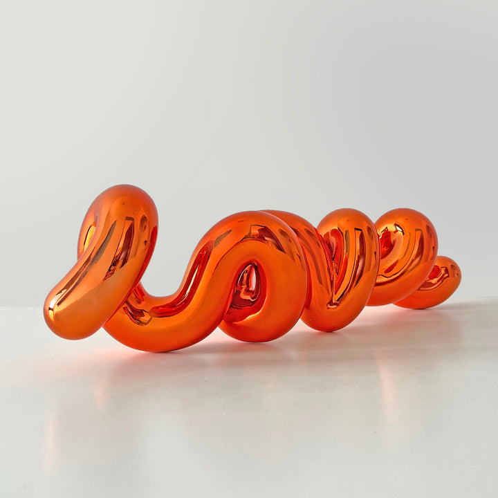 Freestanding sculptural artwork spelling ‘LOVE’ in a flowing, cursive form, finished in high-gloss orange-tinted stainless steel.