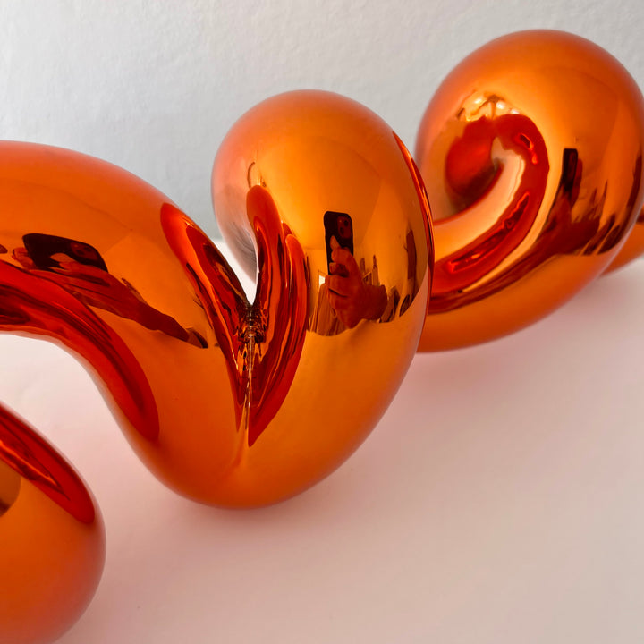 Freestanding sculptural artwork spelling ‘LOVE’ in a flowing, cursive form, finished in high-gloss orange-tinted stainless steel.
