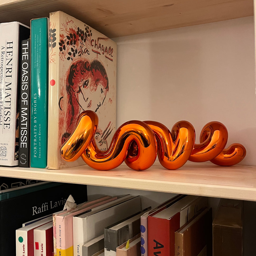Freestanding sculptural artwork spelling ‘LOVE’ in a flowing, cursive form, finished in high-gloss orange-tinted stainless steel.