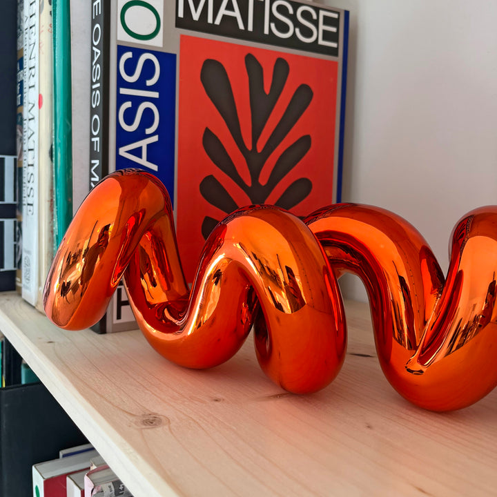 Freestanding sculptural artwork spelling ‘LOVE’ in a flowing, cursive form, finished in high-gloss orange-tinted stainless steel.