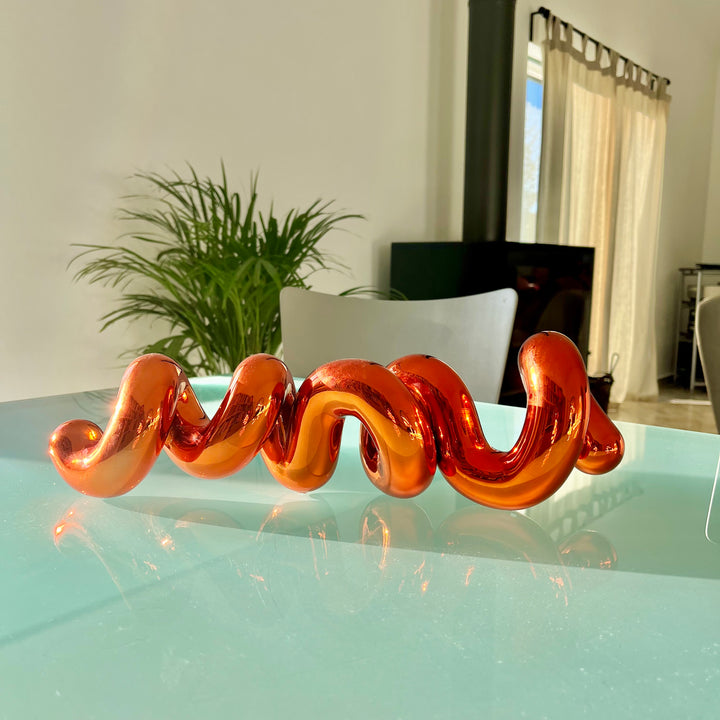 Freestanding sculptural artwork spelling ‘LOVE’ in a flowing, cursive form, finished in high-gloss orange-tinted stainless steel.