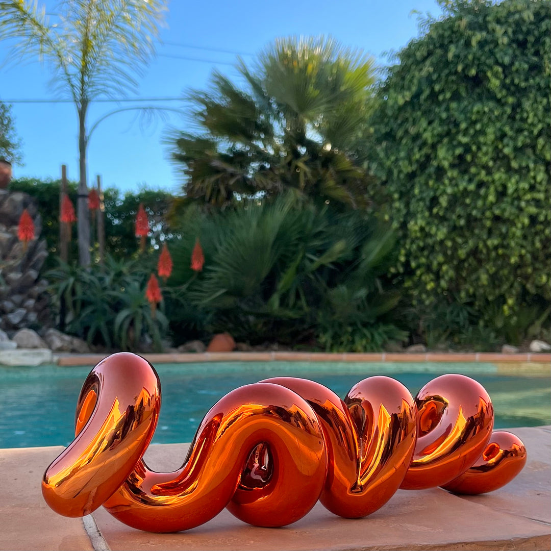 Freestanding sculptural artwork spelling ‘LOVE’ in a flowing, cursive form, finished in high-gloss orange-tinted stainless steel.