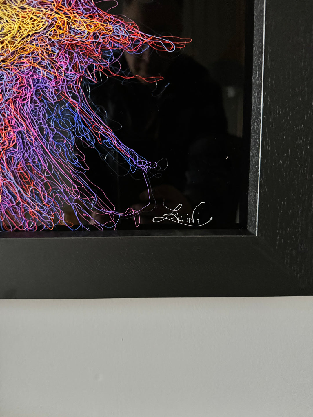 A vibrant textured artwork of a baby orangutan leaning over shimmering water, created with Zalini’s intricate injected-scribble technique, featuring flowing multicoloured lines, bright orange fur, vivid purples and blues, and a mirrored reflection across a glossy black surface.