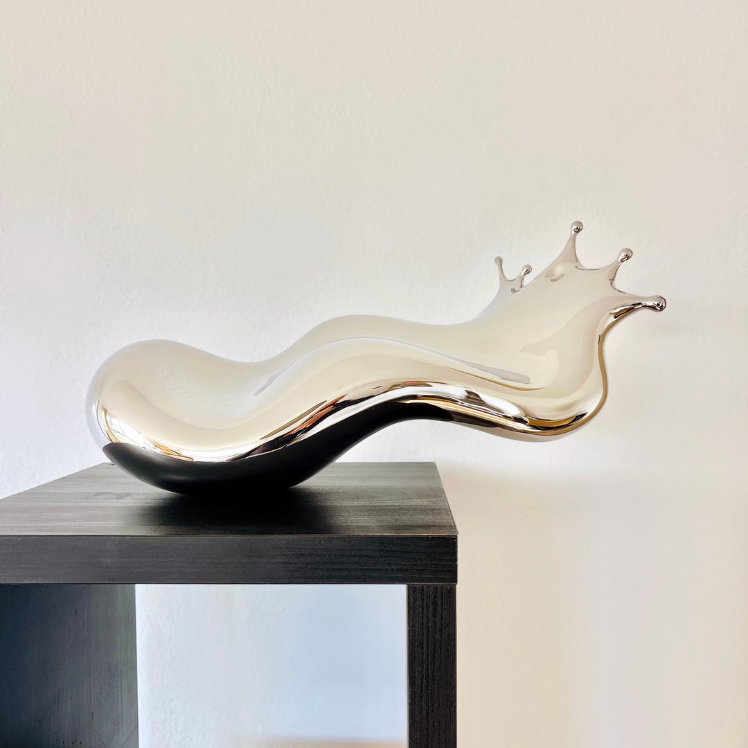 Polished stainless steel sculpture by Yoni Alter capturing the elegant moment of a liquid droplet mid-bounce. The reflective form twists fluidly to the left, with raised crown-like peaks suggesting impact and motion. A sculptural meditation on impermanence and form, grounded by a hidden counterweight for stability.