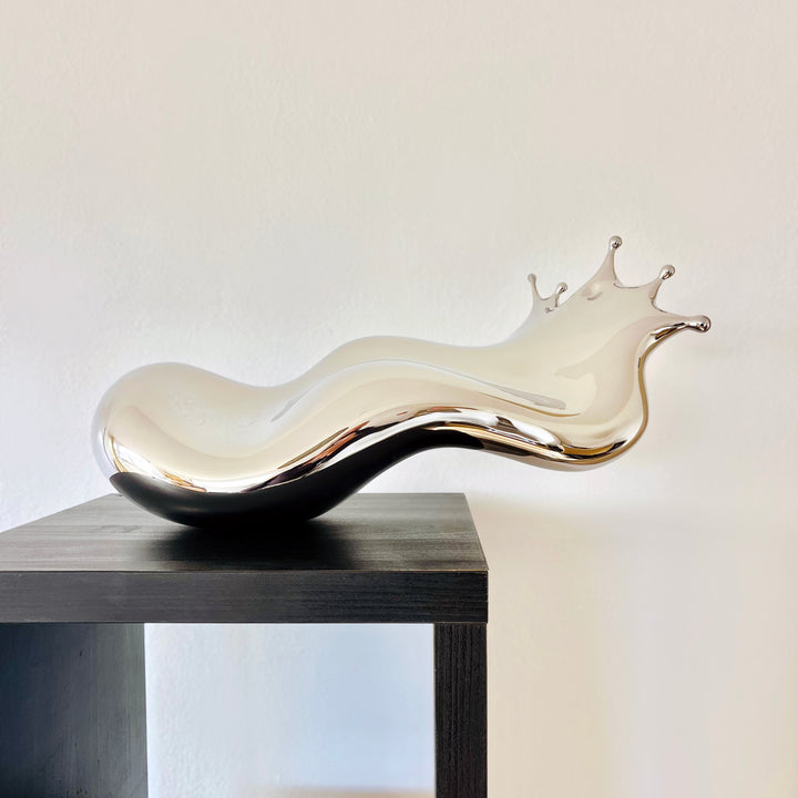Polished stainless steel sculpture by Yoni Alter capturing the elegant moment of a liquid droplet mid-bounce. The reflective form twists fluidly to the left, with raised crown-like peaks suggesting impact and motion. A sculptural meditation on impermanence and form, grounded by a hidden counterweight for stability.